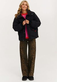 Žena v černé prošívané bundě přes růžový a červený svetr s kalhotami s leopardím vzorem a černými botami, stojící na jednobarevném pozadí.