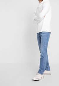 Camicia bianca a maniche lunghe con colletto, jeans azzurri a gamba dritta e sneaker bianche con dettagli rossi, in piedi contro uno sfondo semplice.