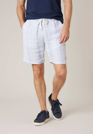 Homme portant un short gris clair à motifs avec un cordon blanc, un t-shirt bleu marine et des baskets bleu marine avec des semelles blanches.