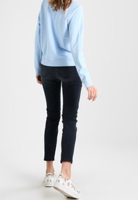 Žena v světle modrém svršku, černých upnutých džínách a bílých teniskách s potiskem zvířecího vzoru, odchází na jednobarevném bílém pozadí.