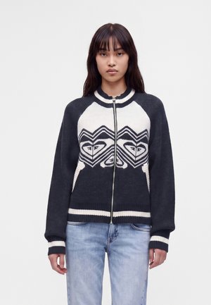 Jonge vrouw die een zwart-witte sweater met rits draagt met hartvormige geometrische patronen, gecombineerd met lichtblauwe spijkerbroek.