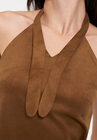 Gros plan d'une personne portant un top en daim marron à col halter en forme de V et larges bretelles nouées à l'avant.