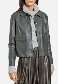 Veste en cuir vert foncé, cropped, avec un col pointu, un système de fermeture à bouton-pression à l'avant et deux poches plaquées. Portée par-dessus un pull à col roulé gris.