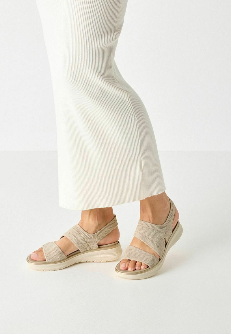 Pieds portant des sandales à brides en tissu beige avec semelles épaisses, associées à un pantalon blanc large en côte sur un fond clair uni.