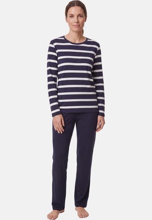 Frau steht barfuß, trägt ein marineblau-weiß gestreiftes Langarmshirt und einfarbige marineblaue Hose vor schlichtem Hintergrund.