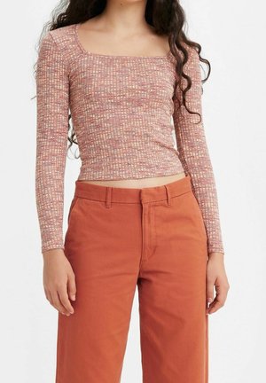 Kobieta ubrana w różową bluzkę z długim rękawem i kwadratowym dekoltem w wzory oraz w wysokie, rdzawe spodnie, stojąca na jednolitym tle.
