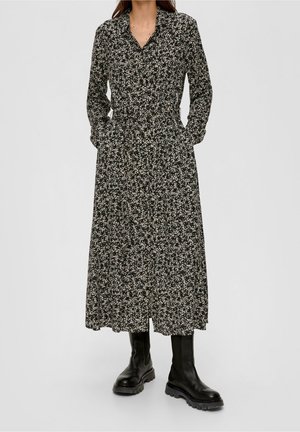 Kvinde iført en langærmet sort og hvid blomster maxi kjole kombineret med sorte chunky læderstøvler, stående på en ensfarvet baggrund.