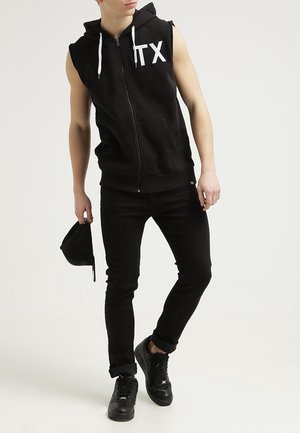 Black sleeveless zip-up hoodie with white lettering, paired with black jeans and black sneakers. The look is completed with a black cap.