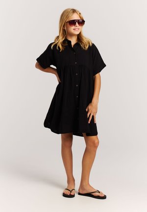 Juoda su sagutėmis užsegama suknelė trumpomis rankovėmis ir sluoksniuotu sijonu, pagaminta iš lengvos medžiagos, derinama su tamsiais akiniais nuo saulės ir šlepetėmis.