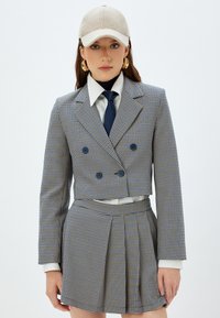 Geknipte dubbelbreasted blazer en geplooide rok in een blauw-geel ruitpatroon, gecombineerd met een witte shirt, stropdas en beige pet.