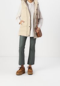 Personne portant un gilet matelassé beige par-dessus une chemise blanche, un pantalon vert court, des chaussures à plateforme marron et portant un sac à bandoulière marron.
