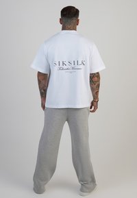 Biała koszulka z krótkim rękawem z czarnym logo "SikSilk" i napisem na plecach, w połączeniu z jasnoszarymi spodniami dresowymi. Gładka faktura materiału.