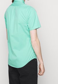 Košile s krátkým rukávem v světlé mátové zelené barvě z bavlny, s plisováním na zádech, zakřiveným lemem a klasickým límcem. Nosí se s černými kalhotami.