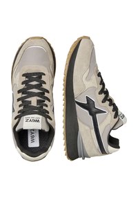 Beige a šedé semišové tenisky s černými akcenty a texturovanými podrážkami. Obsahují detaily loga a černé šňůrky na vzorovaném povrchu.