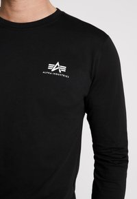 Schwarzes Langarm-Shirt mit rundem Ausschnitt, aus Baumwolle. Mit einem weißen Logo "ALPHA INDUSTRIES" auf der linken Brust.