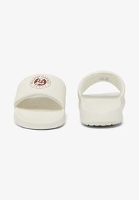 White slide sandals made of smooth material, featuring a rectangular front opening, textured insole, and a circular logo on the strap.