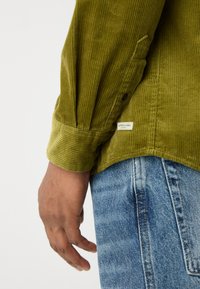 Cămașă de corduroy verde măsliniu cu manșetă cu nasture, având dungi verticale și o etichetă mică a brandului pe tiv. Asortată cu blugi din denim albastru deschis.