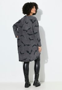 Donna con capelli ricci, che indossa un lungo abito grigio scuro con stampe di pipistrelli neri, leggins in pelle nera e stivali neri con borchie, in piedi di lato.