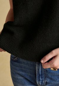 Person richtet den Saum eines schwarzen Strickpullovers, kombiniert mit blauen Jeans, vor einem neutralen Hintergrund.