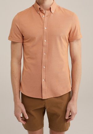 Chemise orange à manches courtes avec un tissu texturé et un petit logo brodé sur la poitrine, accompagnée de shorts marron.
