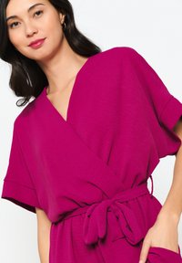 Fuchsia wikkeljurk met korte mouwen, een V-halslijn en een gestrikt tailleband. Zachte, gestructureerde stof met een vloeiende uitstraling.