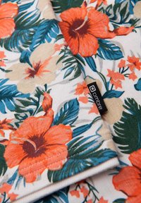 Blommande tyg med orange hibiskus och gröna blad på en ljus bakgrund, med en svart etikett med vit text. Mjuk textur.