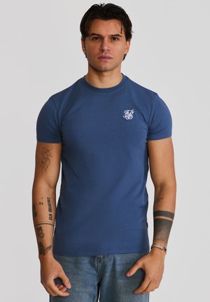 Mann mit dunklem Haar trägt ein eng anliegendes blaues T-Shirt und Jeans, zeigt verschiedene Tätowierungen auf beiden Armen vor einfachem Hintergrund.