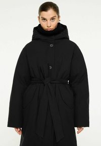 Svart, lång, vadderad kappa med huva, midjebandet och stora framfickor. Slät textur med knappstängningar och hög krage.