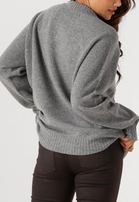 Frau trägt einen lockeren grauen Strickpullover mit langen Ärmeln und dunkle eng anliegende Hosen, steht mit dem Rücken zur Kamera und eine Hand in die Hüfte gestützt.