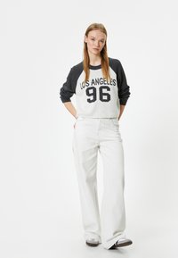 Weißes Sweatshirt mit grauen Raglanärmeln, das "LOS ANGELES 96" in Schwarz zeigt. Kombiniert mit weißen, weit geschnittenen Hosen und grauem Schuhwerk.