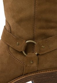 Gros plan d'une botte en daim marron avec une bride décorative et un anneau en métal au-dessus de la semelle cousue.