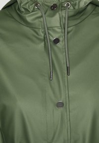 Oliwkowy płaszcz przeciwdeszczowy z wysokim kołnierzem, zatrzaskami, regulowanymi sznurkami oraz gładką, lekko błyszczącą teksturą.