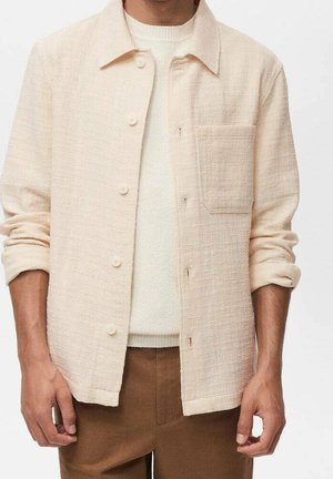 Man wearing a cream textured button-up shirt with rolled sleeves over a white sweater and brown pants, standing against a plain background.