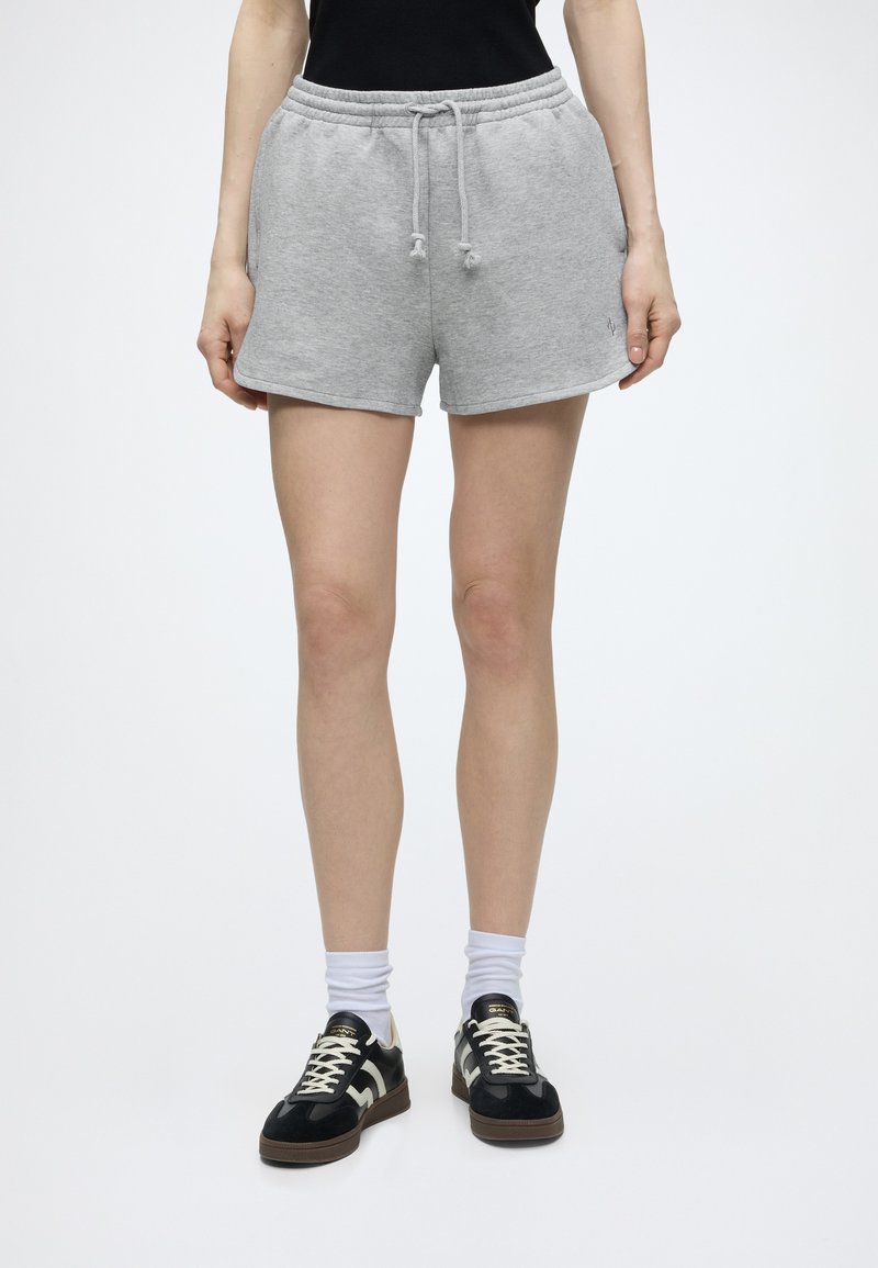 Personne portant un short gris clair à cordon, des chaussettes blanches et des baskets noires avec des lacets blancs, debout devant un fond uni.