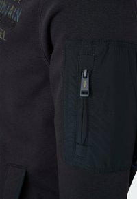Black sweatshirt featuring a smooth texture, standard fit, and a zippered utility pocket with a yellow accent on the left sleeve.