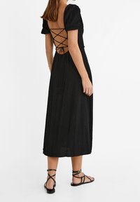 Femme portant une robe midi noire texturée avec des manches bouffantes et un dos noué croisillons, associée à des sandales plates noires à lanières.