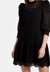 Svart spetsklänning med puffärmar, med en scalloped fåll och intrikata hålmönster över hela. Mjuk textur, figurnära bodice.