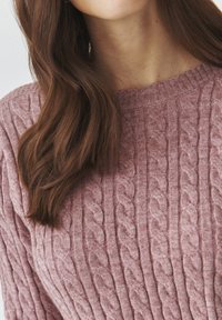 Cable-knit sweater in soft, pale pink wool with a ribbed neck, featuring textured patterns and a cozy appearance.