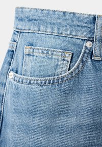 Gros plan d'un jean en denim bleu clair montrant la poche avant et la petite poche pour pièces de monnaie avec des coutures visibles et des rivets métalliques.