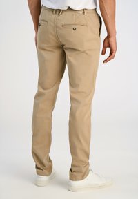 Hombre vistiendo pantalones chinos beige con bolsillos traseros y zapatillas blancas, de pie contra un fondo claro y liso.