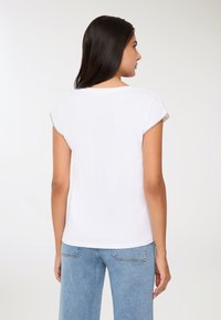 Kobieta z długimi ciemnymi włosami, ubrana w białą bluzkę z krótkim rękawem z ozdobnymi brzegami rękawów oraz jasnoniebieskie dżinsy, odwrócona plecami.