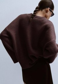 Nadrozměrný tmavě hnědý pletený svetr s texturovaným povrchem, kulatým výstřihem a spadnutými rukávy, doplněný těsně padnoucí černou sukní.