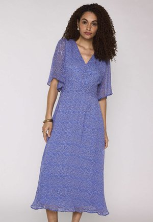 Femme aux cheveux bouclés portant une robe midi bleue à manches courtes, col en V et à petits pois, debout devant un fond uni clair.