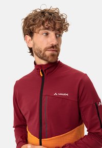 Bordeauxfarbene Softshell-Jacke mit hohem Kragen, kontrastierendem orangefarbenem Taillenbereich, vertikaler Tasche und dem VAUDE-Logo auf der Brust.