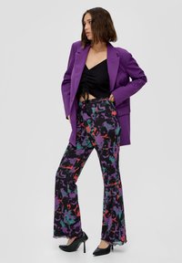 Lila Blazer über einem schwarzen Crop-Top, kombiniert mit ausgestellten Hosen mit einem mehrfarbigen abstrakten Muster. Schwarze High Heels runden das Outfit ab.