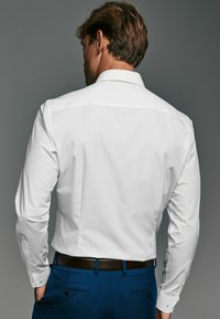 Cămașă albă cu mânecă lungă și guler standard, combinată cu pantaloni albaștri și o curea maro, prezentând o textură netedă și un design clasic.