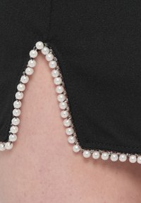 Zwart kledingstuk met een gestructureerde afwerking, met een zoom die wordt geaccentueerd door een rij witte parels, wat een opvallende rand creëert.