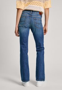 Jeans de mezclilla azules con corte acampanado, que presentan bolsillos traseros y una etiqueta de cuero marrón. La tela tiene un ligero desvanecimiento y se combina con zapatillas blancas.