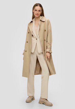 Beige trenchcoat met gouden knopen, lange mouwen en een riem. Daaronder een beige pak met een V-hals top en op maat gemaakte broek. Chunky beige schoenen.