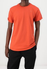 Kortärmad t-shirt med rund hals i livlig orange bomull, med uppvridna ärmar och en liten logotypdetalj på bröstet. Kombinerad med svarta byxor.
